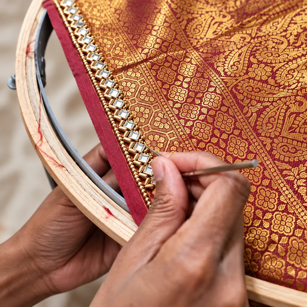 Close-up of SACH Couture hand-finishing a stonework border on maroon and gold brocade fabric.